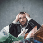 Stressed man in suit holding head surrounded by piles of oversized foreign currency